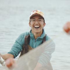 A person stands in the ocean in waders, smiling as they hold a net in the air.