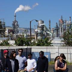 UCLA Law students and faculty standing and walking in front of an energy plant full of machinery and smokestacks, with a blue sky in the background during a toxic tour of Wilmington, CA, a predominantly Latinx neighborhood surrounded by industry and oil.