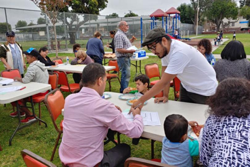 Many people in sitting at tables in a park eating, talking and writing. Jose leans over the table in the foreground pointing to the paper a man is writing on. 