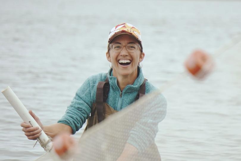 A person stands in the ocean in waders, smiling as they hold a net in the air.