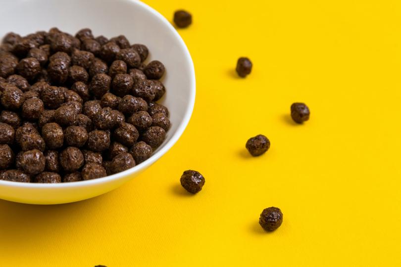 White bowl with chocolate corn cereal balls and milk on yellow background, top-down view.