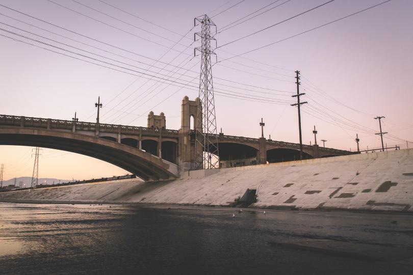Los Angeles River with concrete sides, a bridge and energy pylons surrounding it against a dusk sky.