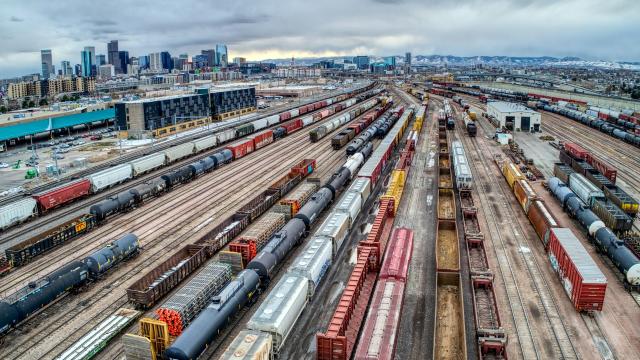 Aerial view of a trainyard with many train tracks, some full of different kinds of trains and others empty. The Denver, CO skyline is in the background under a grey cloudy sky.