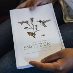 Hands hold a white notebook with an illustration and Switzer Foundation logo on it.