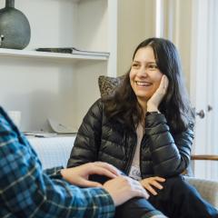 A fellow is sitting, smiling and listening to another person speak.