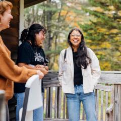 Three people are standing on an outdoor deck, laughing. There are fall leaves in the background. 