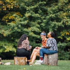 Three Switzer Fellows sit together, laughing, on tree stumps on a grassy lawn with trees in the background.