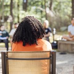 The back of a person's head, with curly, black hair and an orange shirt, sitting in a outdoor circle of people sitting in chairs, with trees and sunlight in the background.