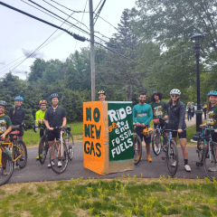 A group of cyclists stand with their bikes, smiling. A green and orange sign in the middle reads 'no new gas' and 'ride to end fossil fuels'.