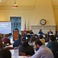 Four speakers sit at a table at the front of a room with a powerpoint and a full audience of people