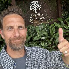 Dave Kramer takes a selfie giving a thumbs up in front of a sign for the Medelin botanical garden.