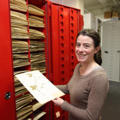 Erika Krimmel stands in front of a large red cabinet with a label that reads "BOTANY". She is smiling at the camera and holding a preserved botanical specimen: a pressed plant on a white paper.