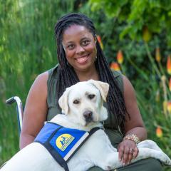 India Harville sits in a wheelchair with her white Labrador service dog, Nico, on her lap. They are outside with green plants in the background.