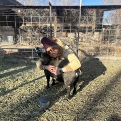 Maya Higgins crouches, wearing a maroon winter hat, hugging a small grey and black goat on her home farm. There are farm structures and plants in the background.