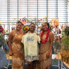 Mica and Maya stand with another person in front of a display of flowers and clothing, holding a tie dye t-shirt that reads "NO WAR, NO ICE, HELIX FOR LIFE, New Haven CT, April 17 2026" in blue text.