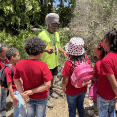 Students in red t-shirts stand listening to a man in a yellow shirt and knitted hat speaking outside at the St. George Village Botanical Garden as he discusses the slave grave site located within the gardens.