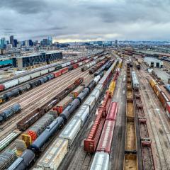 Aerial view of a trainyard with many train tracks, some full of different kinds of trains and others empty. The Denver, CO skyline is in the background under a grey cloudy sky.