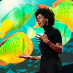 Ayana Elizabeth Johnson is seen on stage from below, with a screen showing colorful fish on it as the background. She is wearing a microphone, a black short-sleeved turtleneck and holding both hands out as she speaks.