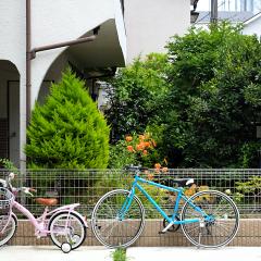 A pink child's bike and a blue adult's bike rest on a fence near concrete buildings and manicured green trees. 