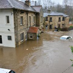 Brown water floods a street in Ireland. A white car is mostly submerged in the water, and the water is halfway up the doors to the buildings on the street. The buildings are made of stone bricks with tile roofs. 