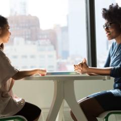 Two women sitting at a tall table next to windows with city buildings in the distance.