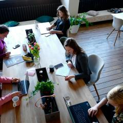 Seen from above: five women sit around a wooden table with laptops and notebooks, smiling and working together.
