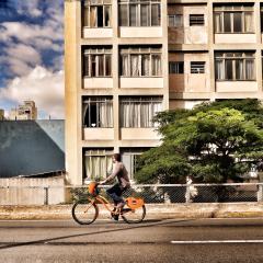 A person is biking on a road with a tree and a concrete apartment building in the background.