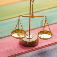 Golden scales with a wooden base rest on a wooden surface that is painted in colorful stripes. 