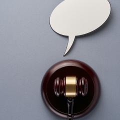 A brown legal gavel sits in a holder on a grey background. An empty white speech bubble sits above it.