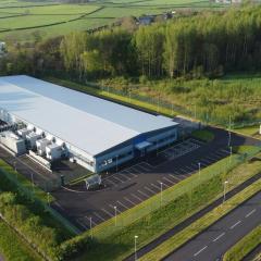 An aerial view of a Data Centre in Coleraine, Ireland. It’s a large white building with a small parking lot. There are small roads, green grass, woods, and a farm in the surrounding area. 