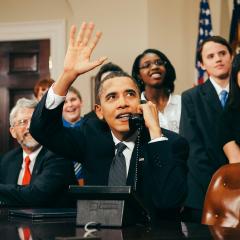 President Obama sits at a desk in the White House, holding a black phone and waving while looking up and to the side. Senators sit with him and middle school students stand behind them.