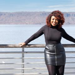 Janelle is smiling and standing in front of a railing with water and hills in the background