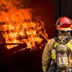 A firefighter stands facing away from the camera, toward a structure burning with bright orange flames