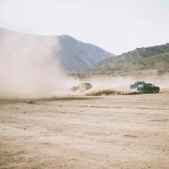 Two jeeps and a car drive on a dusty flat landscape with green hills in the background, kicking up a large dust cloud.