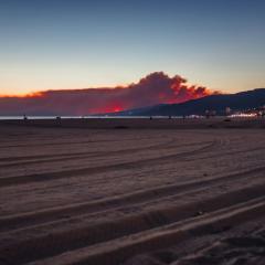 A ground level photo shows beach sand in the foreground, with a coastline and hills with city lights at dusk in the background, with a huge cloud of grey wildfire smoke backlit with the orange of the sunset.