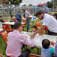 Many people in sitting at tables in a park eating, talking and writing. Jose leans over the table in the foreground pointing to the paper a man is writing on. 