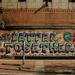 A mural is spray painted onto a building that is boarded up with plywood. The mural reads BETTER TOGETHER! with hands holding each other, a globe, red hearts, and a city skyline.  