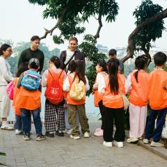 Children wear orange vests on a field trip outside with four adults. They are standing in a courtyard near a body of water and green plants.