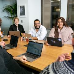 A group of people sit around a table with laptops open at a meeting, smiling and laughing with each other.