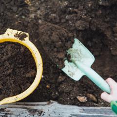 A child's hand scoops brown soil into a yellow pot with a gardening trowel. 