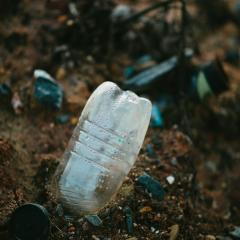 A plastic water bottle pokes out of brown soil dotted with rocks, with other plastic trash out of focus in the background. 