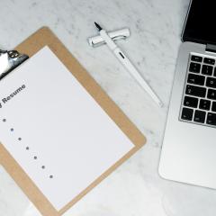 A clipboard with a resume template, a pen and a mac laptop sit on a white marble table.