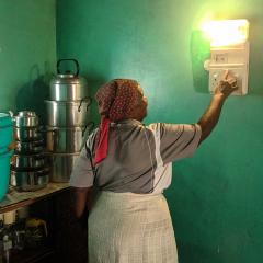 An African woman in a headscarf and apron faces away, turning on an electric wall lamp mounted on a green wall in a kitchen. Pots and pans are on a counter behind her and the profile of another person's face is watching and speaking, cut off on the left.