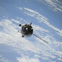A satellite in orbit, seen from above against a backdrop of clouds and ocean on earth.