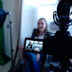 A woman is being filmed by a video recording camera, seen from behind the camera.