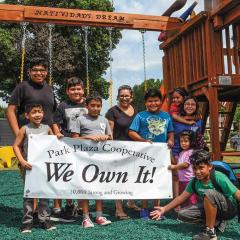 A group of children and adults stand in front of a playground holding a white banner that says "We Own It!"