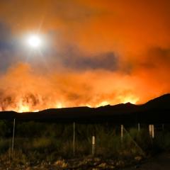 Fires burn over the crest of a dark hill with orange smoke rising.