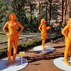 Life-sized, bright orange statues of three women, including Dr. Kimberley Miner, in a brick courtyard outside a brick Smithsonian building with trees in the background.