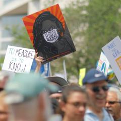 At a climate march, a hand holds up a sign of showing a Black woman against an orange background, wearing a white mask that reads PEOPLES CLIMATE MARCH.