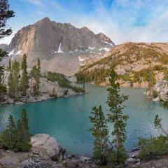 Pine trees and rocks surround a turquoise blue lake with a mountain and blue sky in the background. 